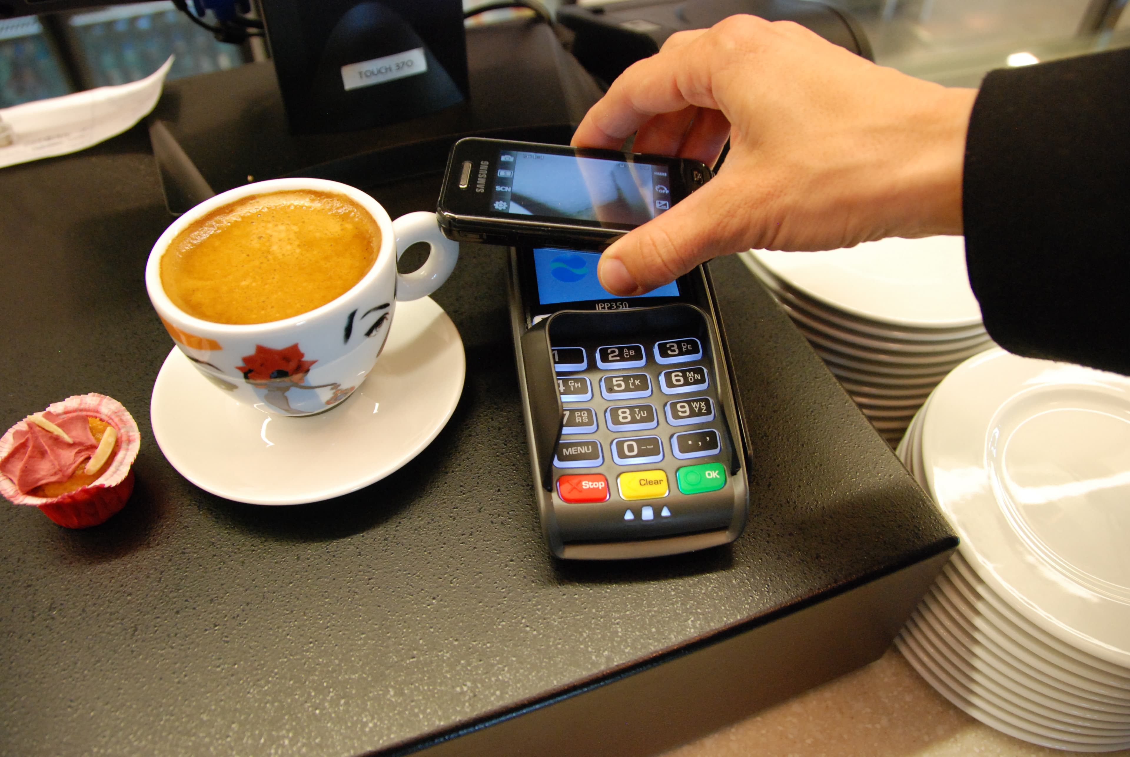 A hand holding a mobile phone above a payment terminal beside a coffee cup